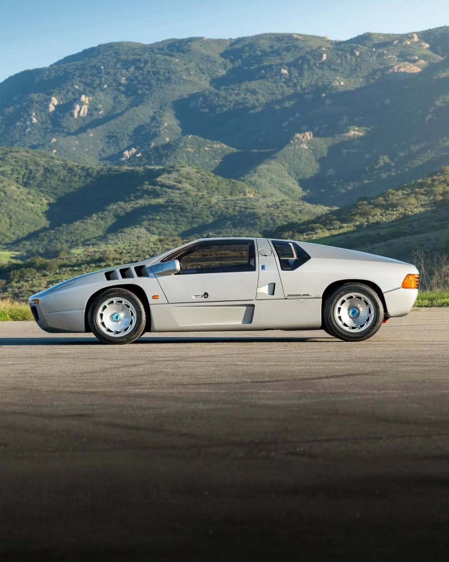 نمای جانبی ابرخودروی کلاسیک ایزدرا امپراتور 108i با رنگ نقره‌ای، آینه پیروسکوپی روی سقف و درهای بال‌کبوتری در یک فضای نمایشگاهی.